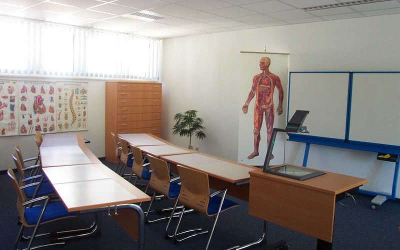 Unterrichtsraum mit Tischen, Stühlen, einer Tafel und einem Schaubild des menschlichen Körpers