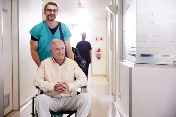 Male Orderly Pushing Senior Male Patient Being Discharged From Hospital In Wheelchair