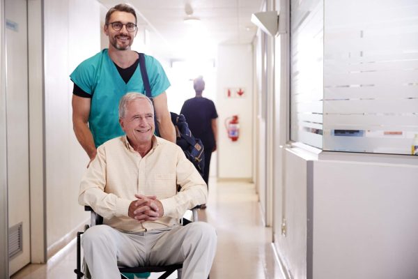 Male Orderly Pushing Senior Male Patient Being Discharged From Hospital In Wheelchair Male Orderly Pushing Senior Male Patient Being Discharged From Hospital In Wheelchair